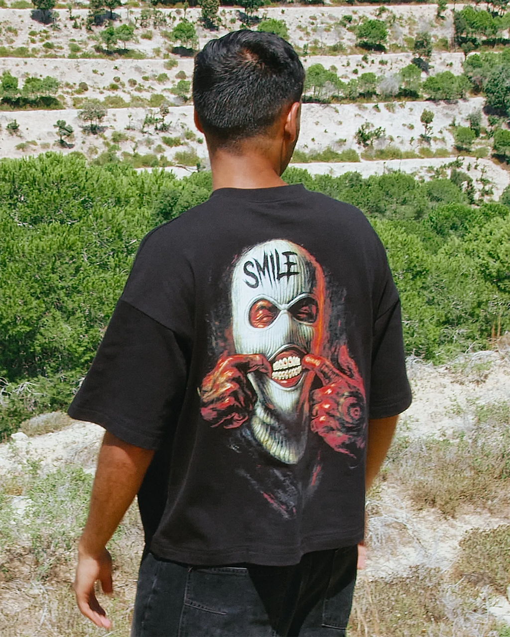 A man viewed from behind wearing a Black graphic t-shirt with a large print of the character Smile on the back, set against a background of green hills and terraced stone walls.