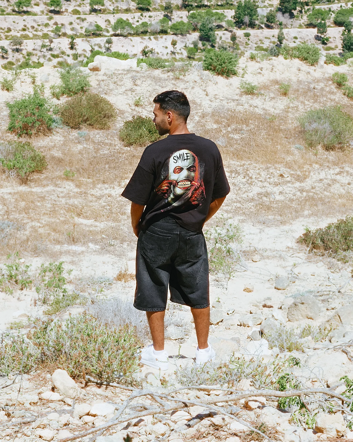 A man stands outdoors in a dry, rocky landscape wearing a Black graphic T-shirt with a horror-style face print and the word 'SMILE' on the back, paired with dark shorts and white sneakers.