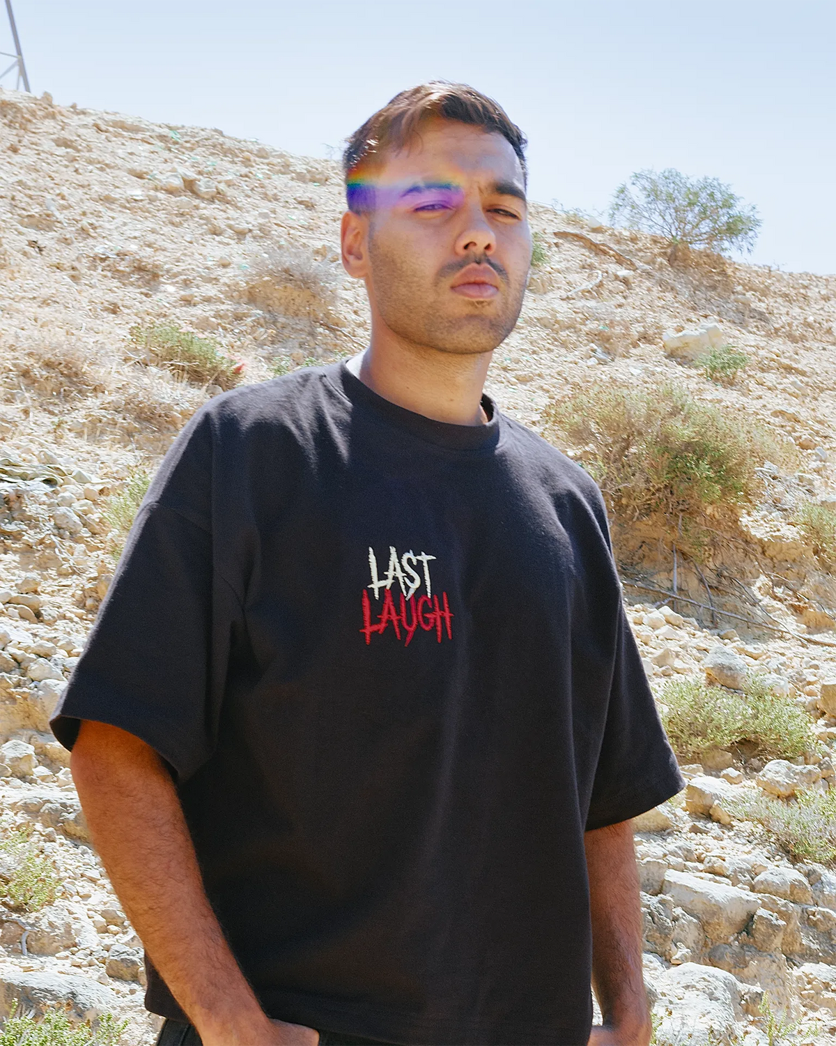 A man wearing a Black oversized t-shirt with 'LAST LAUGH' printed on the chest, standing outdoors against a rocky desert hillside background.