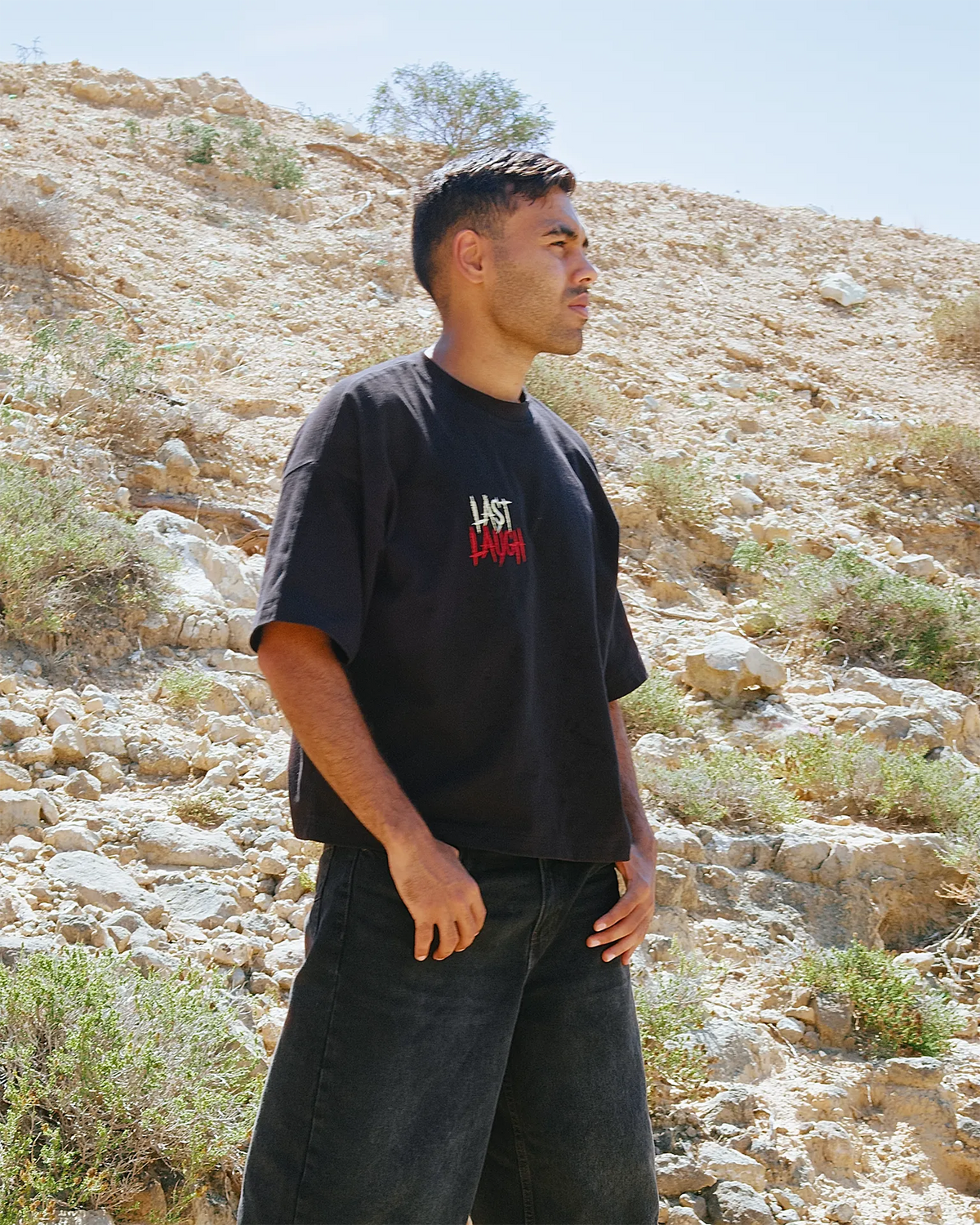 A man wearing a Black oversized t-shirt with 'LAST LAUGH' printed on the chest, standing outdoors against a rocky desert hillside background.