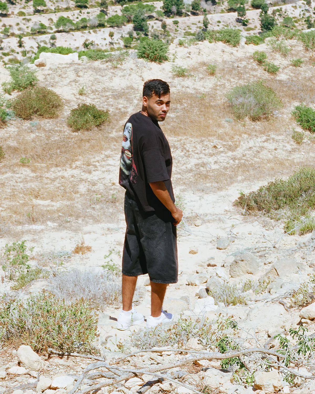 A man stands outdoors in a dry, rocky landscape wearing a Black graphic T-shirt with a horror-style face print and the word 'SMILE' on the back, paired with dark shorts and white sneakers.